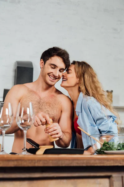 Smiling girl standing near shirtless boyfriend while he seasoning dish — Stock Photo