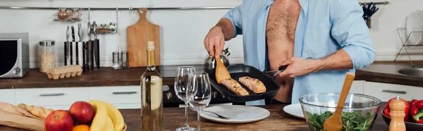 Panoramic shot of man holding frying pan and cooking fish in kitchen — Stock Photo