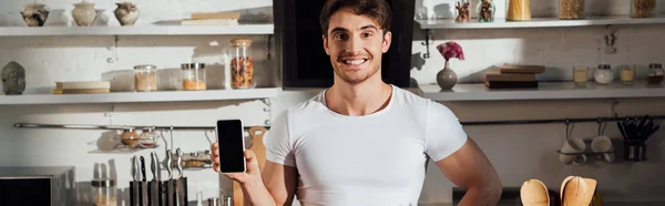 Panoramic shot of smiling muscular man in white t-shirt showing smartphone with blank screen in kitchen — Stock Photo