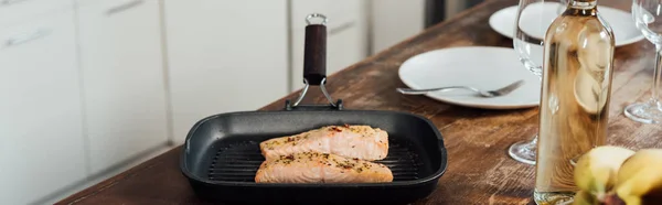 Panoramaaufnahme einer Pfanne mit Fisch, einer Flasche Wein, Weingläsern und Tellern auf dem Tisch in der Küche — Stockfoto