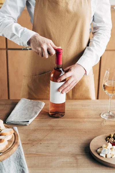 Cropped view of sommelier in apron opening bottle of wine at table — Stock Photo