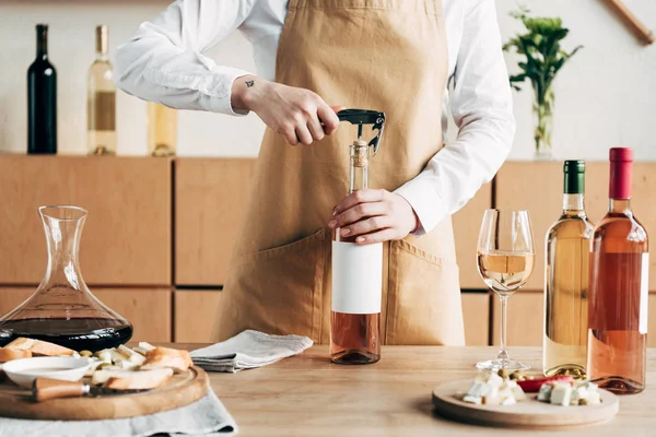 Vista ritagliata del sommelier in grembiule aprendo bottiglia di vino a tavola — Foto stock