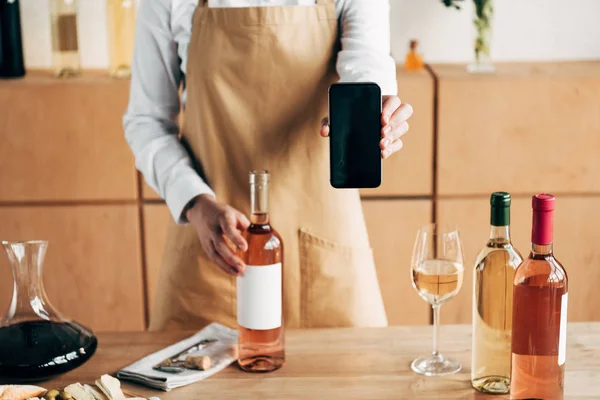 Vista parcial del sommelier en delantal de pie cerca de la mesa con botellas de vino y mostrando el teléfono inteligente con pantalla en blanco - foto de stock