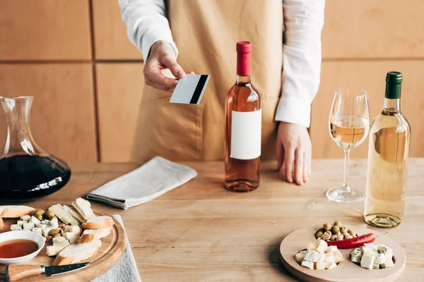 Vista ritagliata del sommelier in grembiule in piedi vicino al tavolo con bottiglie di vino e in possesso di carta di credito — Foto stock