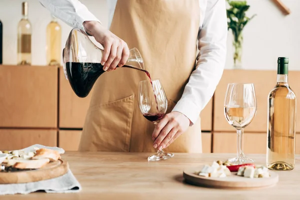 Vista parziale del sommelier in brocca di azienda grembiule e versando vino in bicchiere di vino — Foto stock