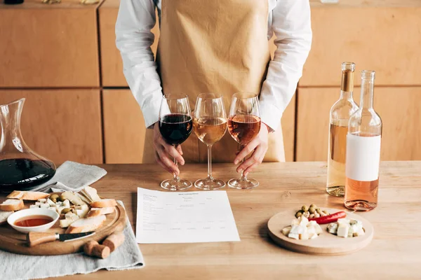 Vista cortada de sommelier em avental segurando copos de vinho perto de mesa de madeira — Fotografia de Stock