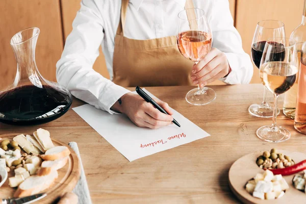 Vista recortada de sommelier en delantal sentado a la mesa, sosteniendo copa de vino y escritura en documento de degustación de vino - foto de stock