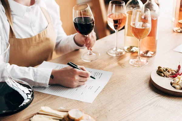 Обрізаний вид сомельє в фартусі, сидячи за столом, тримаючи келих для вина і пишучи в дегустаційному документі — стокове фото