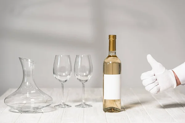 Partial view of waiter in white glove showing thumb up near table with bottle of wine, wine glasses and jug — Stock Photo