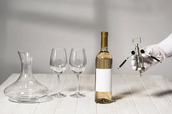 Cropped view of waiter in white glove holding corkscrew near table with bottle of wine, wine glasses and jug — Stock Photo