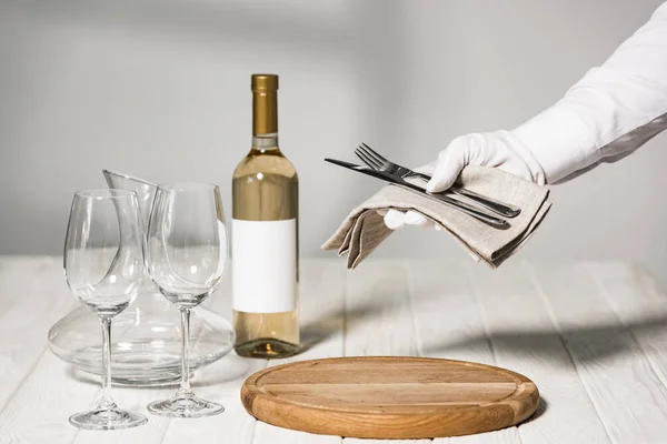 Cropped view of waiter in white glove holding cutlery near table with bottle of wine, wine glasses and jug — Stock Photo