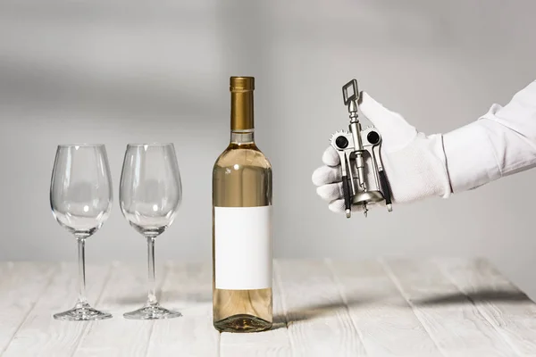 Vista ritagliata del cameriere in guanto bianco con cavatappi vicino al tavolo con bottiglia di vino e bicchieri di vino — Foto stock