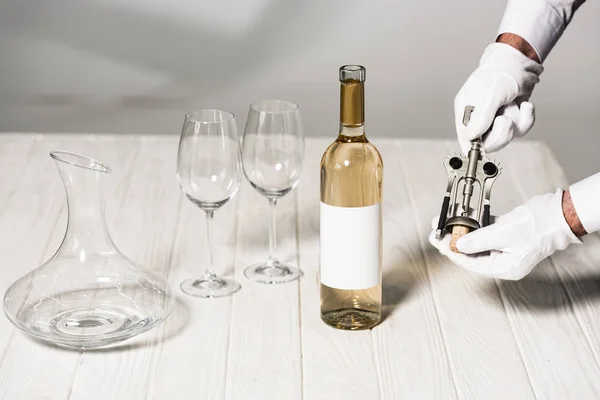 Partial view of waiter in white gloves holding corkscrew on table near bottle, wine glasses and jug — Stock Photo