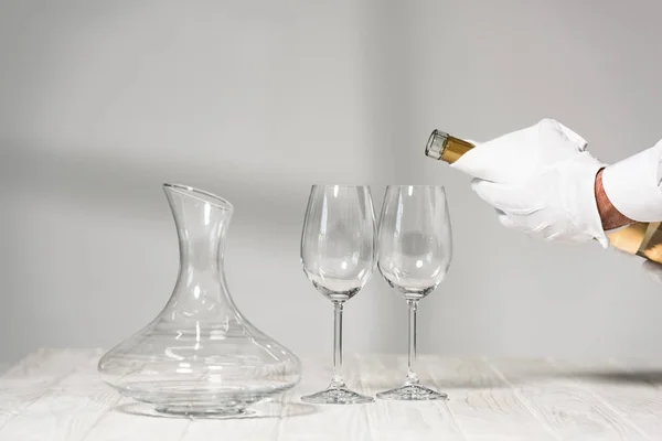 Vista recortada de agua en guante blanco sosteniendo botella de vino cerca de copas de vino en mesa de madera - foto de stock