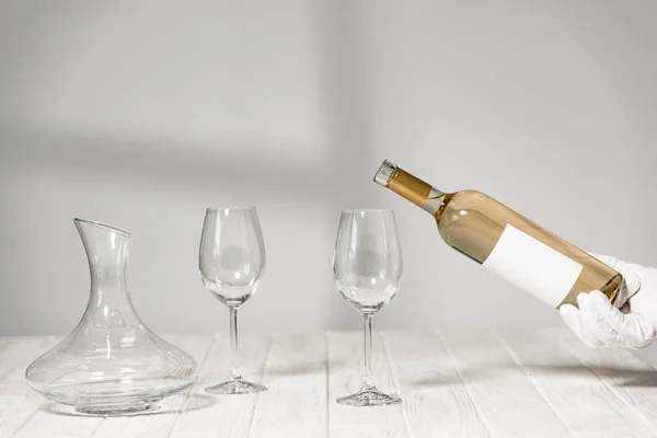 Cropped view of water in white glove holding bottle of wine near wine glasses on wooden table — Stock Photo