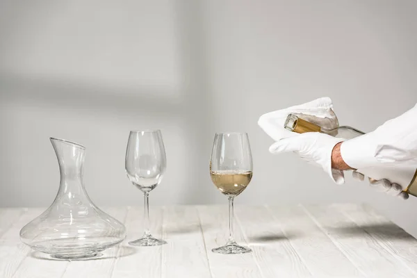 Cropped view of water in white glove holding bottle of wine near wine glasses on wooden table — Stock Photo