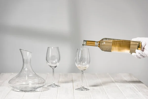 Cropped view of water in white glove holding bottle of wine near wine glasses on wooden table — Stock Photo