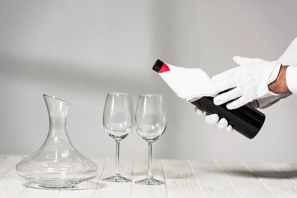 Vue recadrée de l'eau dans des gants blancs tenant une bouteille de vin près de verres à vin sur une table en bois — Photo de stock