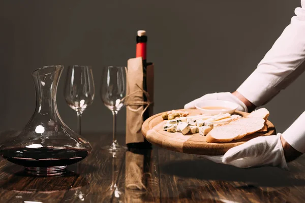 Partial view of waiter in white gloves holding cutting board with cheese and bread near table with wine — Stock Photo