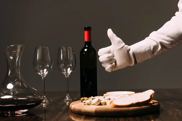 Обрезанный вид официанта в белой перчатке с большим пальцем рядом со столом с едой и вином — стоковое фото