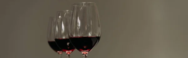 Tiro panorâmico de três copos de vinho com vinho tinto isolado em cinza — Fotografia de Stock