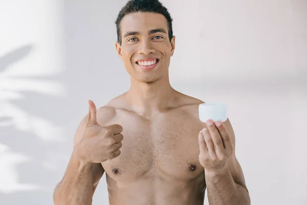 Hombre guapo sin camisa sosteniendo crema cosmética, mostrando el pulgar hacia arriba, sonriendo y mirando a la cámara - foto de stock