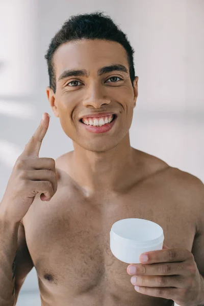 Bonito misto raça homem colocando alguns cosméticos creme com dedo no rosto enquanto segurando recipiente, sorrindo e olhando para a câmera — Fotografia de Stock