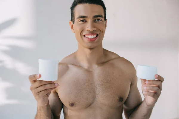 Joven sin camisa sosteniendo dos contenedores para crema cosmética, sonriendo y mirando a la cámara - foto de stock