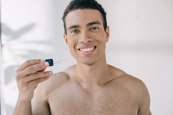 Apuesto hombre de raza mixta sosteniendo la pipeta con suero, sonriendo y mirando a la cámara - foto de stock