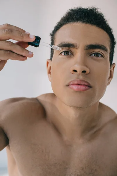 Portrait shot of mixed race man dropping some serum on face with pipette while looking at camera — Stock Photo