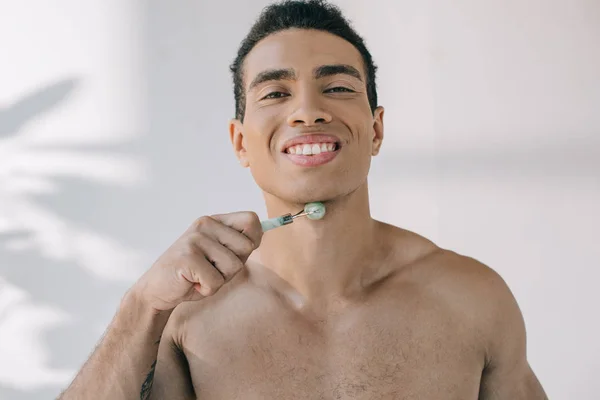 Handsome mixed race man massaging chin with jade roller and smiling while looking at camera — Stock Photo