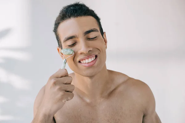 Handsome mixed race man massaging face with stone jade roller and smiling with closed eyes — Stock Photo