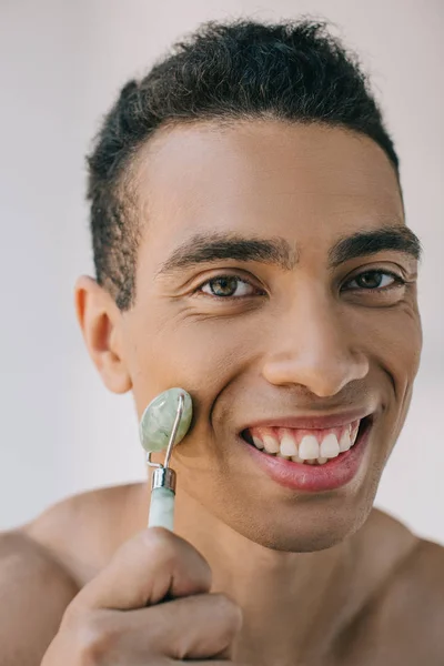 Retrato de hombre guapo masajeando la cara con rodillo de jade de piedra mientras sonríe y mira a la cámara - foto de stock