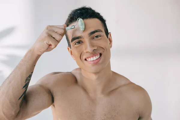 Handsome mixed race man massaging forehead with jade roller and smiling while looking at camera — Stock Photo