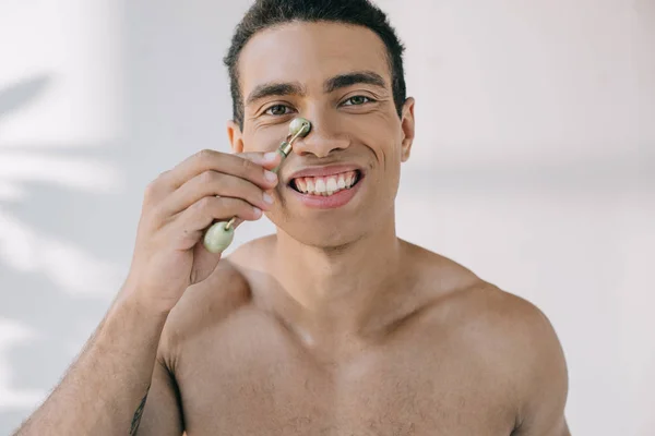 Musculoso hombre de raza mixta masajeando la nariz con rodillo de jade de piedra y sonriendo mientras mira a la cámara - foto de stock