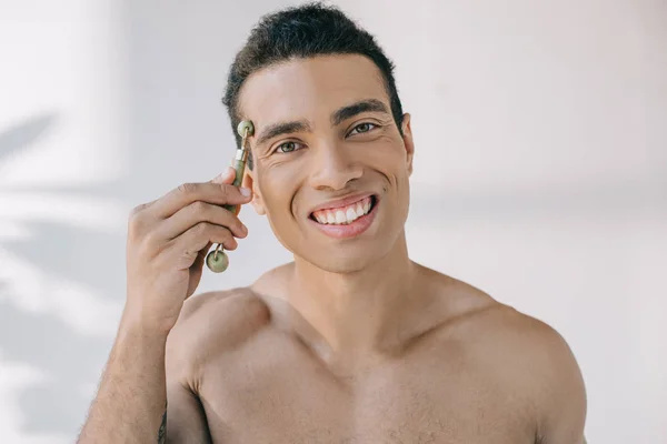 Muscular mixed race man massaging temple with stone jade roller and smiling while looking at camera — Stock Photo