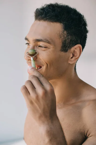 Musculoso hombre de raza mixta masajeando la nariz con rodillo de jade de piedra y sonriendo mientras mira hacia otro lado - foto de stock