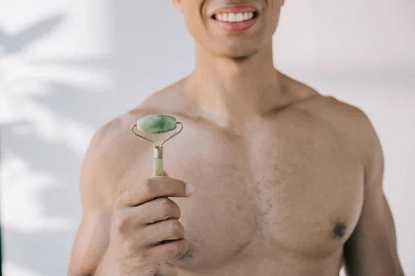 Cropped view of mixed race man presenting stone jade roller and smiling while looking at camera — Stock Photo