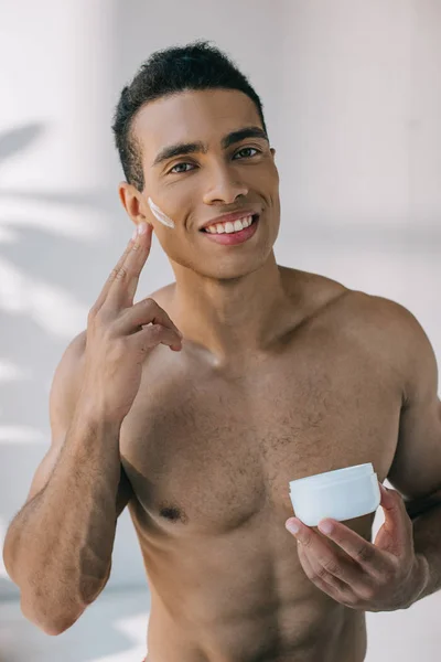 Мускулистый мужчина смешанной расы держит контейнер и наносит косметический крем на лицо, глядя в камеру — стоковое фото
