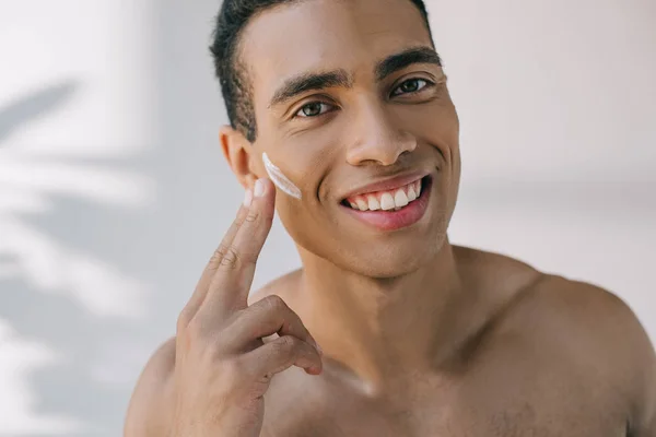 Retrato tiro de homem bonito aplicando creme cosmético no rosto com os dedos e sorrindo enquanto olha para a câmera — Fotografia de Stock