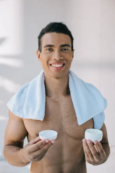Mixed Race junger Mann mit blauem Handtuch auf den Schultern, der einen Behälter mit Kosmetikcreme hält, während er in die Kamera blickt — Stockfoto