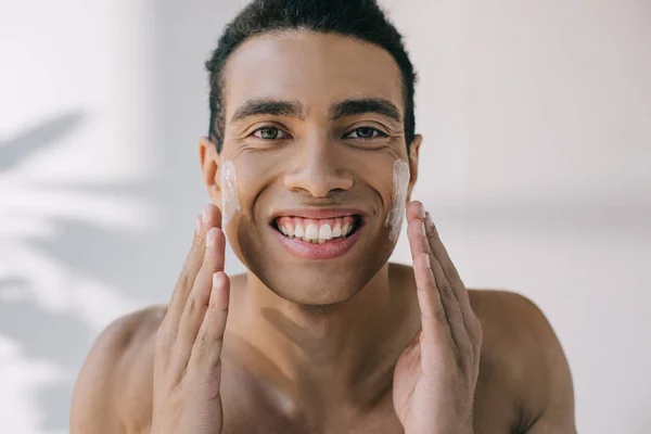 Guapo hombre de raza mixta aplicando crema cosmética en los ckeeks con las manos y sonriendo mientras mira a la cámara - foto de stock