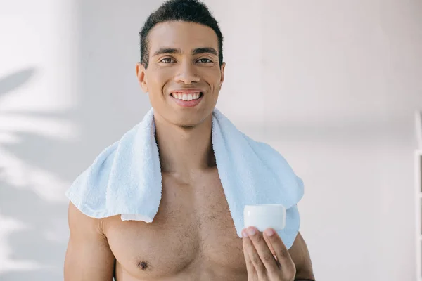 Mixed race young man with blue towel on shoulders holding container with cosmetic cream while looking at camera — Stock Photo