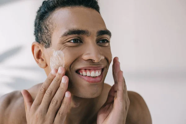 Guapo hombre de raza mixta aplicando crema cosmética en los ckeeks con las manos y sonriendo mientras mira hacia otro lado - foto de stock