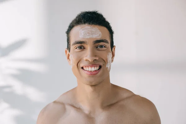 Guapo hombre de raza mixta con crema cosmética en la cara sonriendo mientras mira a la cámara - foto de stock
