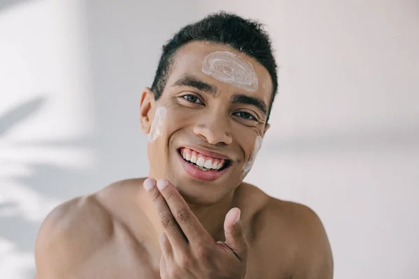 Bonito misto raça homem aplicando creme cosmético no rosto com as mãos e sorrindo enquanto olha para longe — Fotografia de Stock