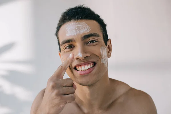 Handsome mixed race man applying cosmetic cream on face with finger and smiling while looking away — Stock Photo