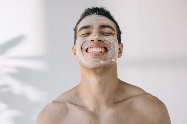 Bonito misto raça homem com cosmético creme no rosto sorrindo com olhos fechados — Fotografia de Stock