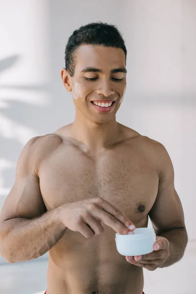 Musculoso hombre de raza mixta tomando crema cosmética con los dedos del contenedor mientras sonríe y mira hacia otro lado - foto de stock