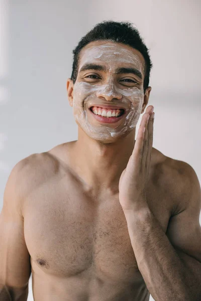 Musculoso hombre de raza mixta aplicando crema cosmética en la cara con la mano y sonriendo mientras mira a la cámara - foto de stock